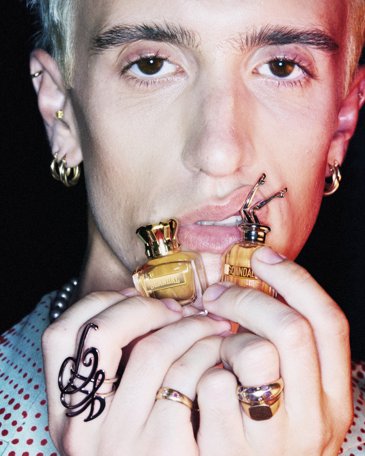Close-up of a blonde adult male holding two bottles of gold perfume in his hand, wearing multiple rings, earrings, and a pearl necklace.