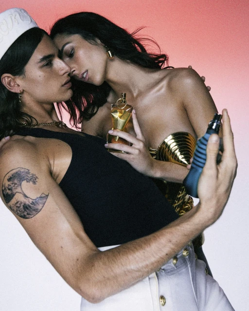 Fragrance ad featuring two models: a man in a sailor cap and a woman in a gold top, both holding fragrance bottles against a gradient background.