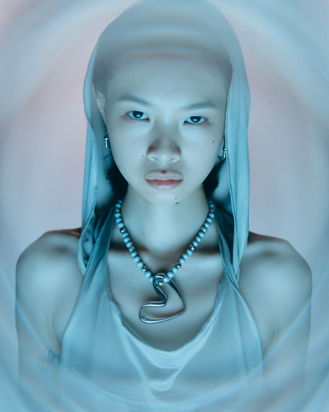Portrait of a young Asian woman wearing beaded necklace with heart pendant and hooded top, gazing intently toward the camera.