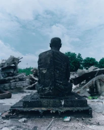 Rear view of a dark textured statue seated amidst concrete rubble under an overcast sky, photographed by Lee Wei Swee.