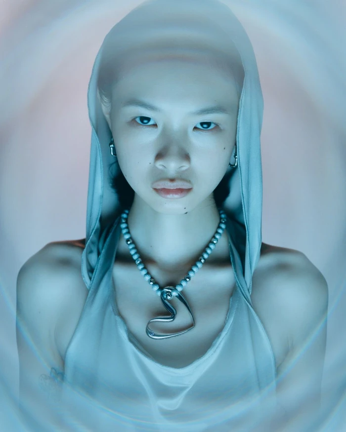 Portrait of a model wearing a hooded grey top and a large heart pendant necklace against a swirling abstract background.