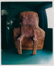A chunky, textured brown terracotta armchair sculpture positioned inside a dark green room with a window and a teal floor.
