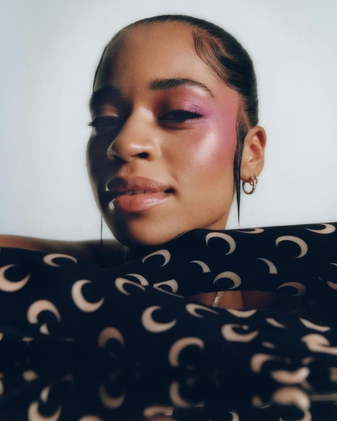 Close-up portrait of a woman with pink eyeshadow and patterned top against a white background in a beauty and fashion setting.