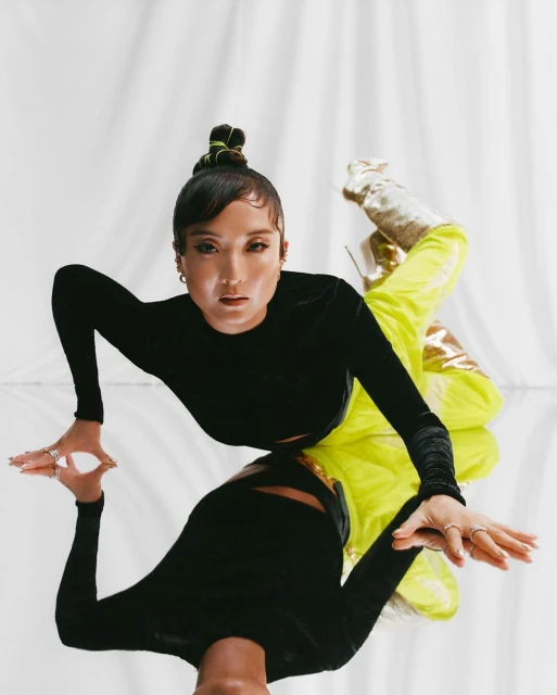 Fashionable adult female with neon yellow pants and gold boots lies on reflective surface in studio with white backdrop.