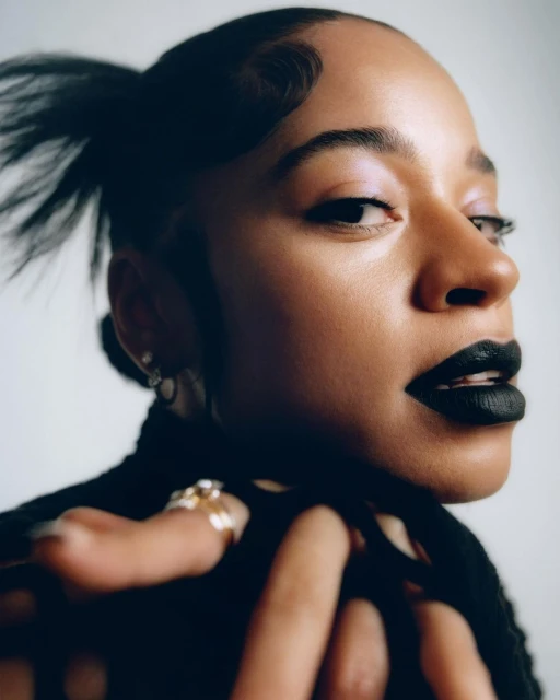 Close-up portrait of an adult woman with dark hair and black lipstick, wearing a black top and rings, posing in a photo studio.