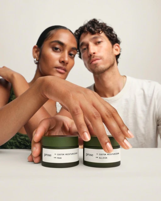 Two skincare products on a table with two adults looking at the camera. The skincare products are Prose custom moisturizers.