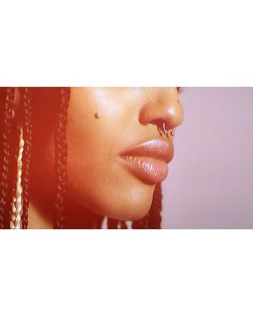 Close-up of an adult female's face featuring a nose ring, glossy lips, and braided hair with blonde strands, captured in a studio.