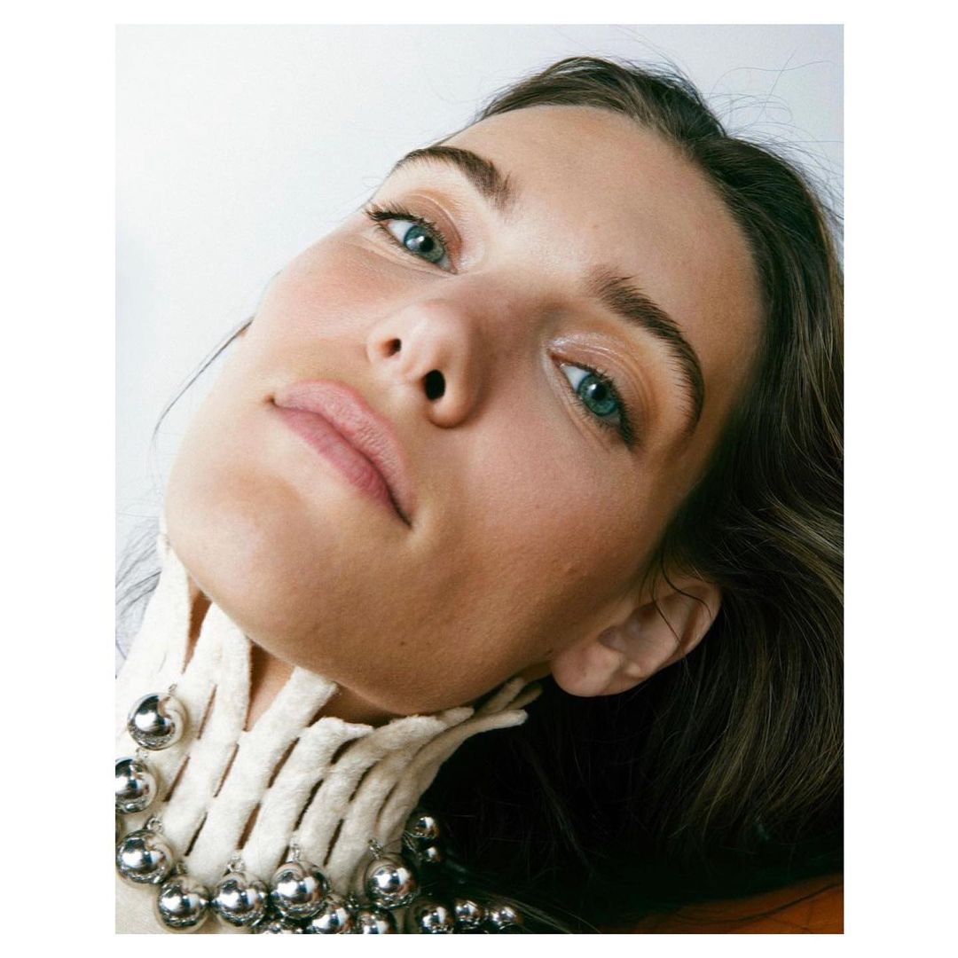Headshot of a young adult female with fair skin and green eyes, wearing an avant-garde white collar and silver bead necklace. Shot indoors.