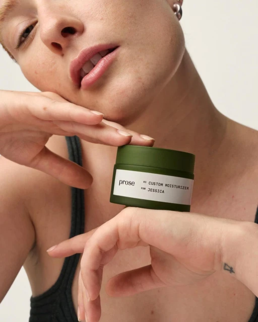 Close-up of a light-skinned adult woman holding a green and white Prose custom moisturizer jar against a light background.