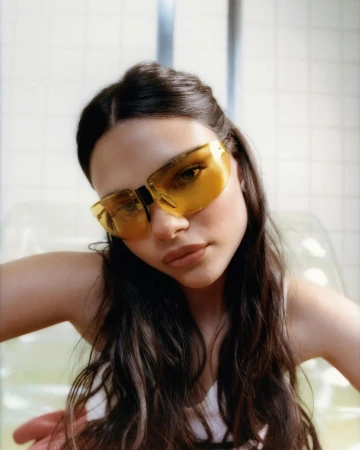 Close-up portrait of a woman wearing tinted yellow sunglasses and a white top against a tiled background.