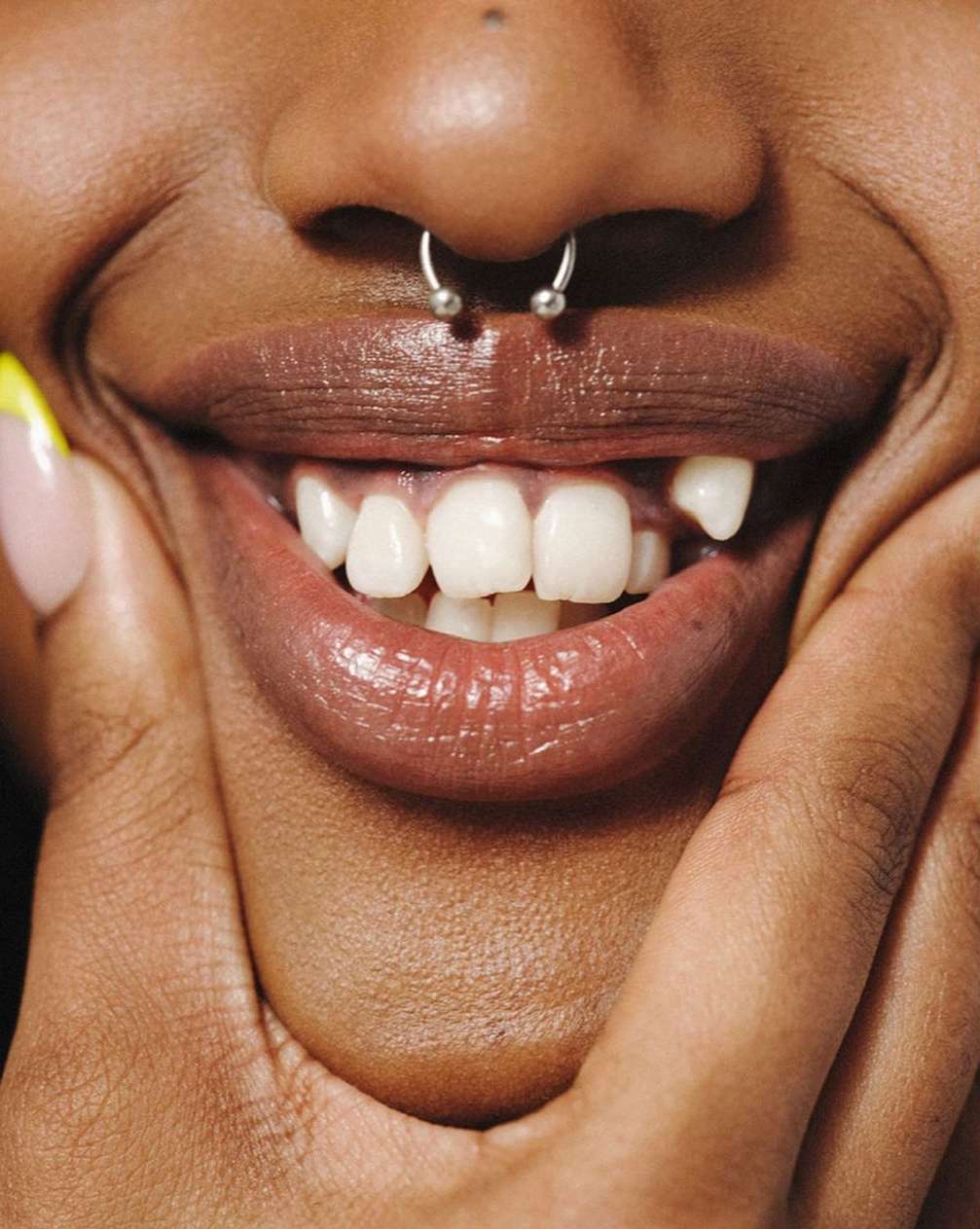 Close-up of a smiling Black woman with a nose ring and glossy lips, showing healthy teeth and a confident expression indoors.