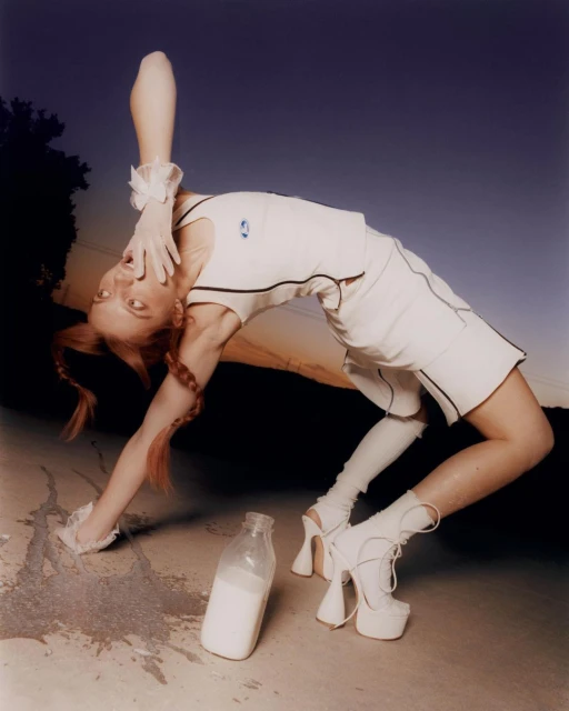 A young adult woman in a white outfit poses in a backbend next to a bottle of milk against a blue sky backdrop, with bare feet and gloves.