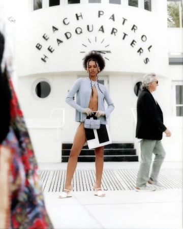 A fashion shoot features a female model in a powder blue cardigan and black-and-white skirt, posing in front of a white building.