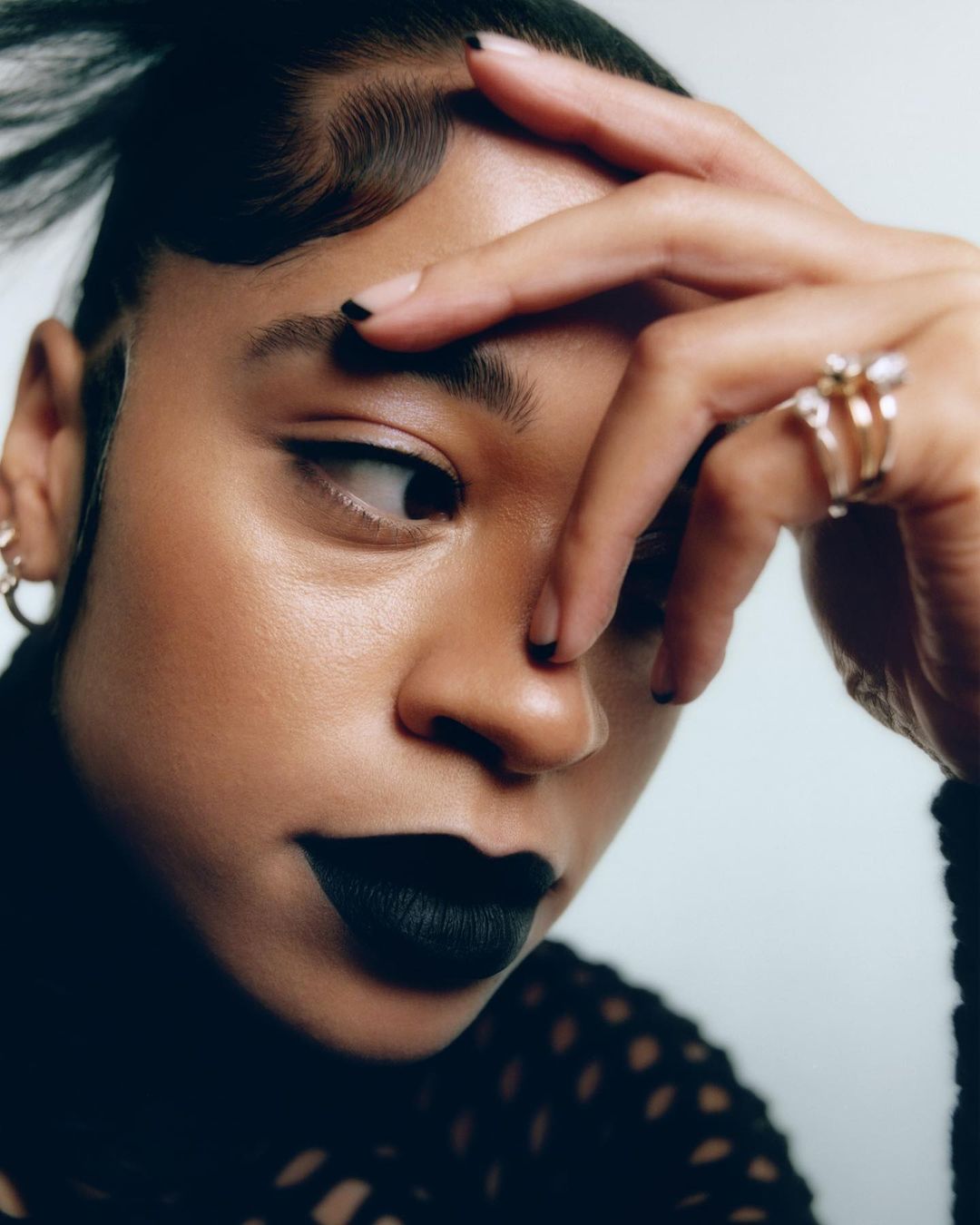 Close-up portrait of a woman with black lipstick and nail polish, wearing a black shirt and gold earrings against a white background.