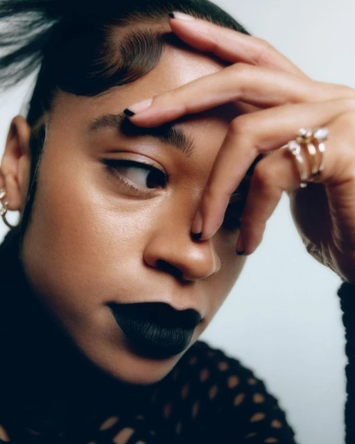 Close-up portrait of a woman with black lipstick and nail polish, wearing a black shirt and gold earrings against a white background.