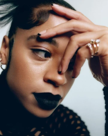 Close-up portrait of a woman with black lipstick and nail polish, wearing a black shirt and gold earrings against a white background.