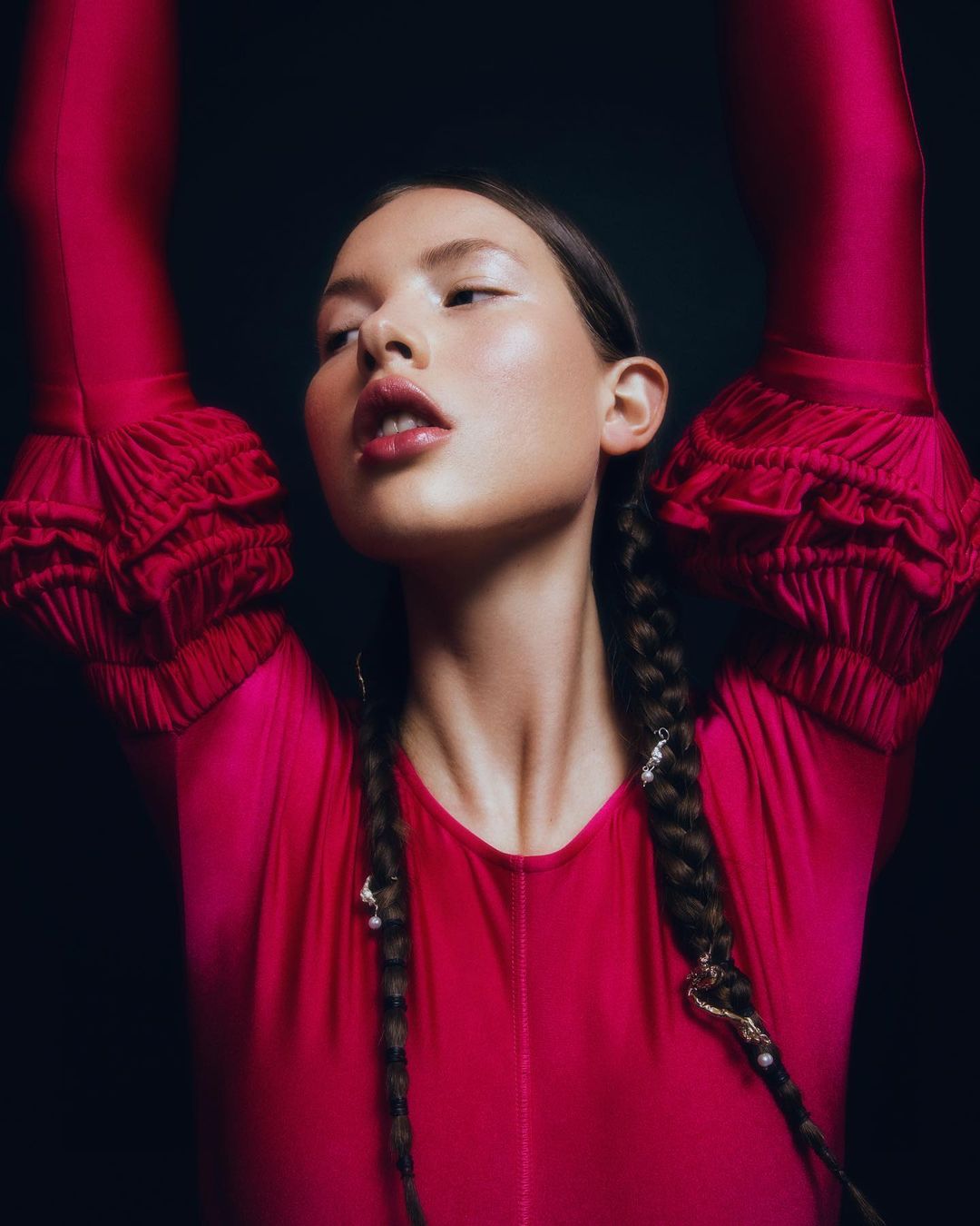Young woman in a red bodysuit poses with arms raised against a dark background, showcasing a fashion or beauty aesthetic.