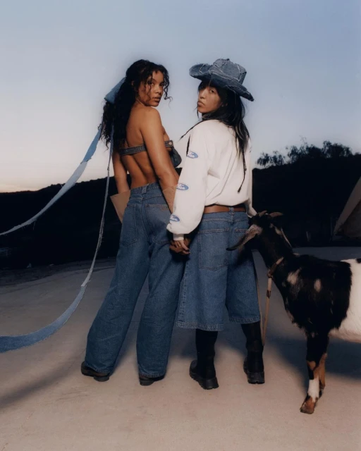 Two women in denim outfits pose with a goat in an outdoor setting against a dusky sky, showcasing casual fashion and rural aesthetics.