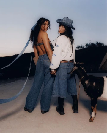 Two women in denim outfits pose with a goat in an outdoor setting against a dusky sky, showcasing casual fashion and rural aesthetics.