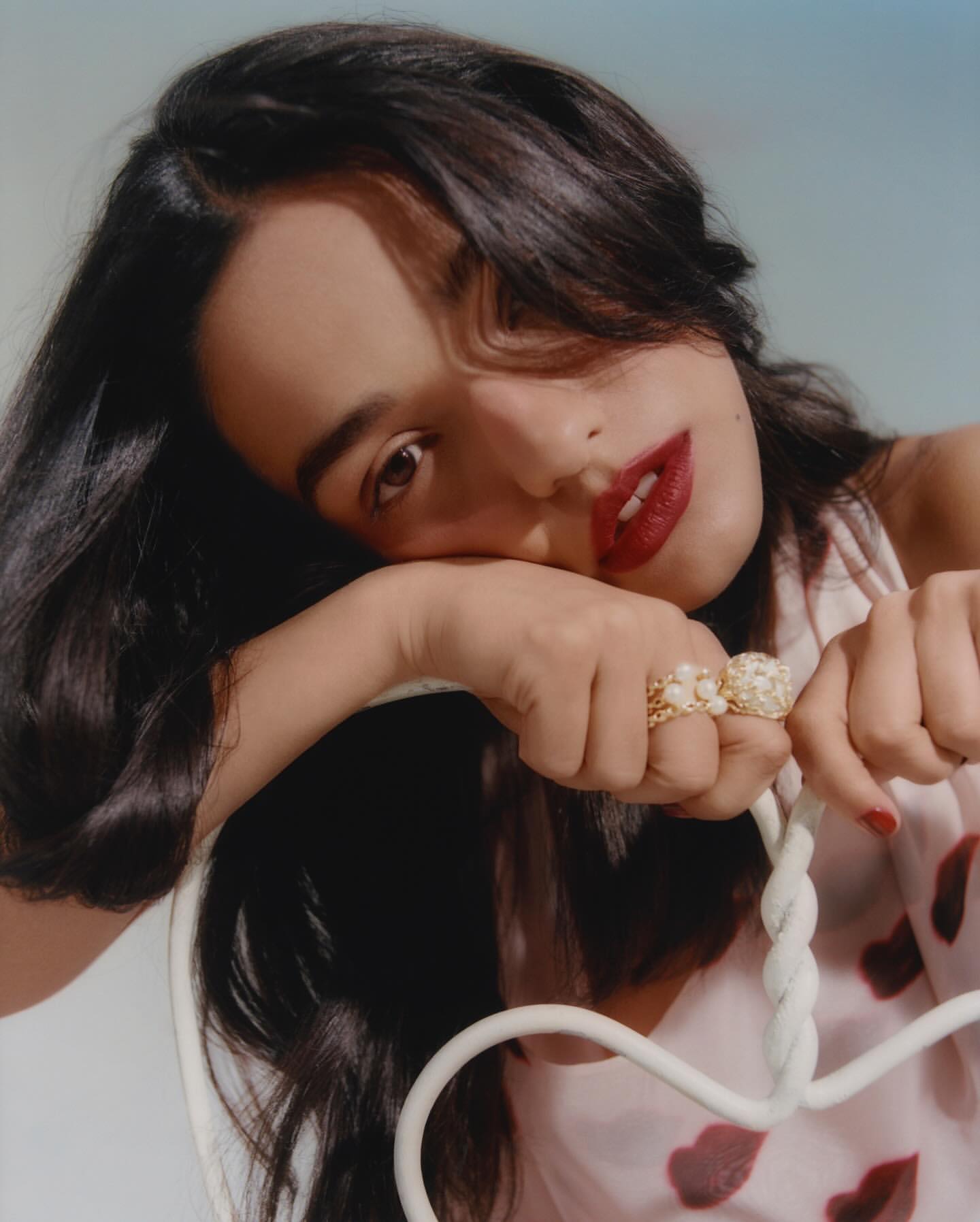Brunette woman with red lipstick rests her head on her hand showcasing large, gold rings in an indoor studio environment with soft lighting.