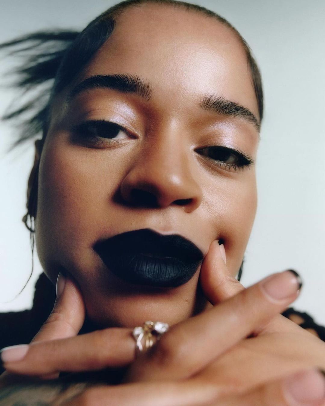 Close-up portrait of a young adult female with dark hair and black lipstick, posing with her hands near her face, wearing a ring.
