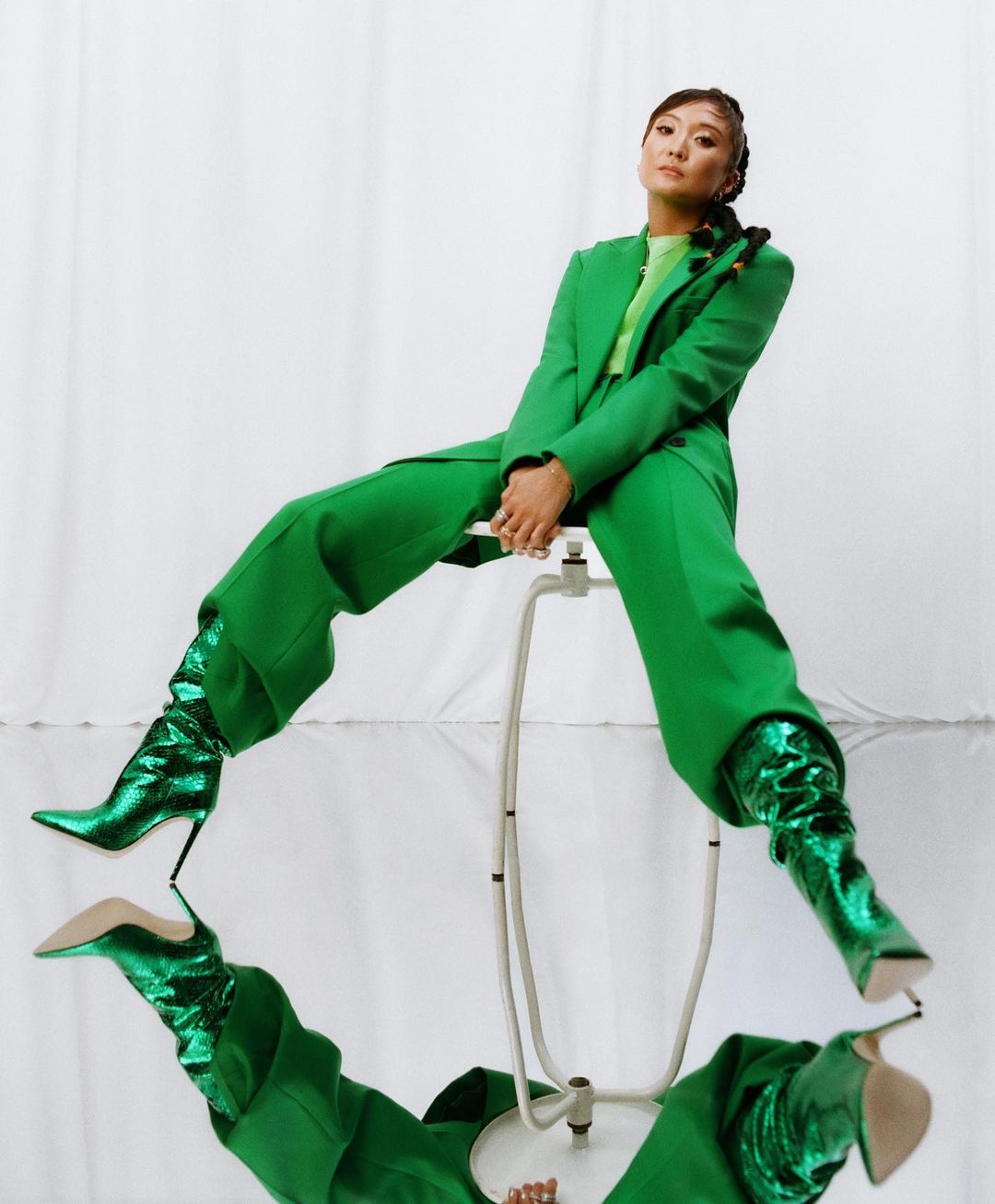 Asian woman in green pantsuit and boots sitting on stool in front of mirror, creating reflection; indoor, studio shot.