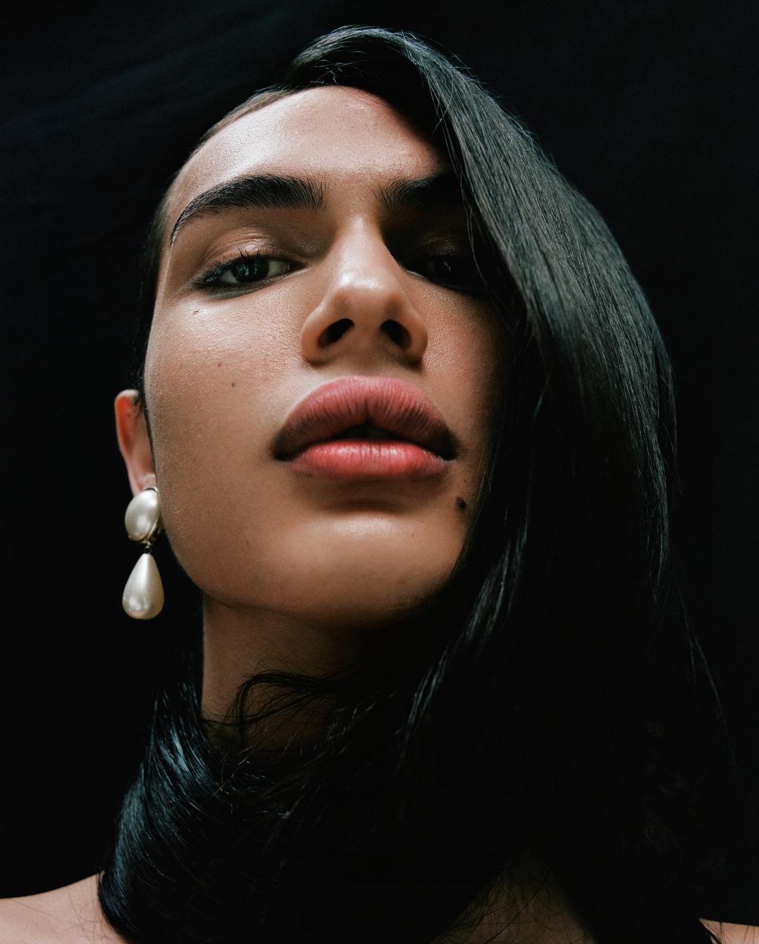 Portrait of a person with dark hair, wearing large pearl earrings, looking up at the camera against a dark, contrasting background.