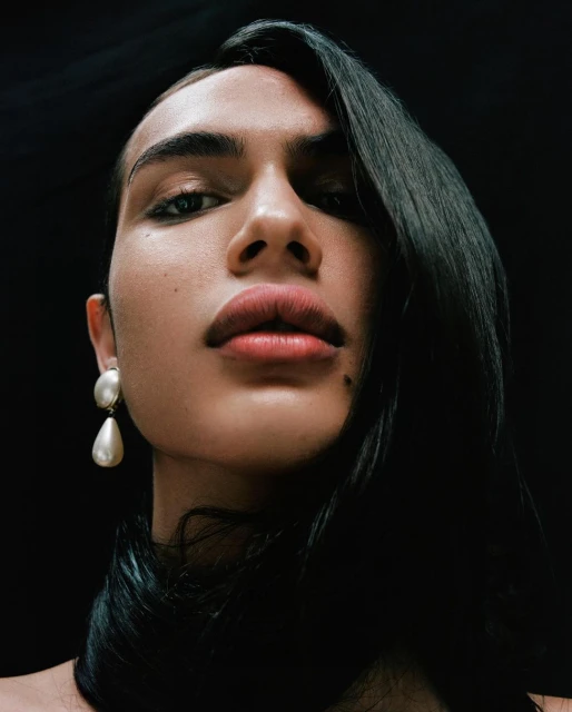 Portrait of a person with dark hair, wearing large pearl earrings, looking up at the camera against a dark, contrasting background.