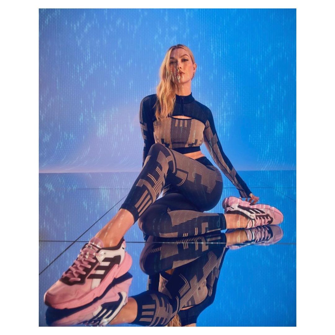 Athletic woman in patterned long-sleeved crop top and leggings poses in pink and black sneakers on reflective surface in studio.