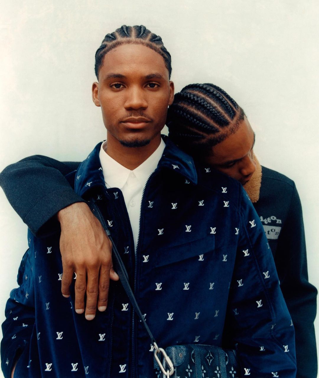 Two adult men in a close studio portrait. One man in a blue jacket with LV logos rests his head on the other man's shoulder.