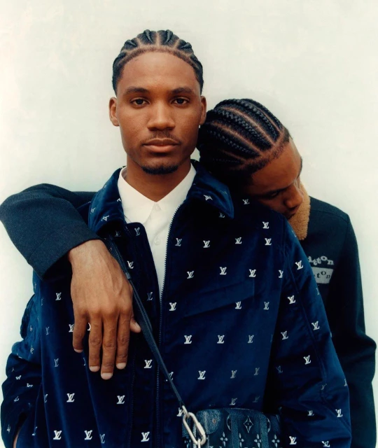 Two adult men in a close studio portrait. One man in a blue jacket with LV logos rests his head on the other man's shoulder.