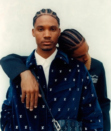 Two adult men in a close studio portrait. One man in a blue jacket with LV logos rests his head on the other man's shoulder.