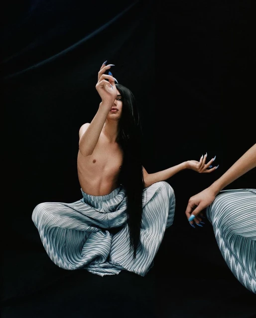 Model with long black hair poses topless in pleated pants with long fingernails against a dark backdrop in a studio shot.