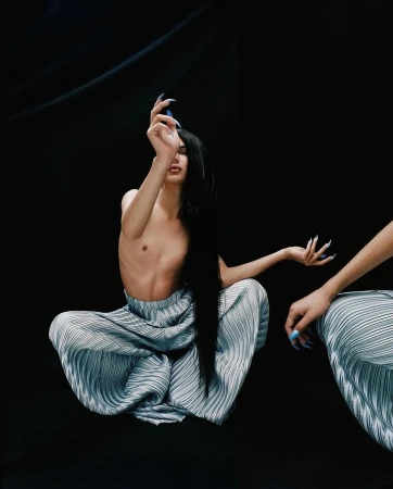 Model with long black hair poses topless in pleated pants with long fingernails against a dark backdrop in a studio shot.