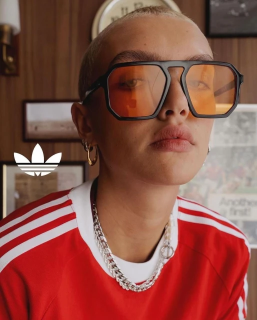Close-up of a short-haired person with a red and white Adidas shirt and glasses in an indoor setting with a wooden background.