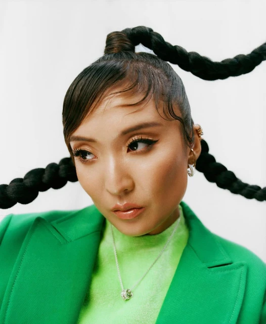 Portrait of a woman with braids, wearing a green blazer and jewelry, against a white backdrop in a close-up studio shot.