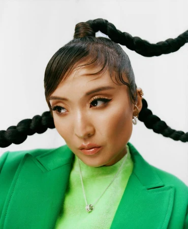 Portrait of a woman with braids, wearing a green blazer and jewelry, against a white backdrop in a close-up studio shot.