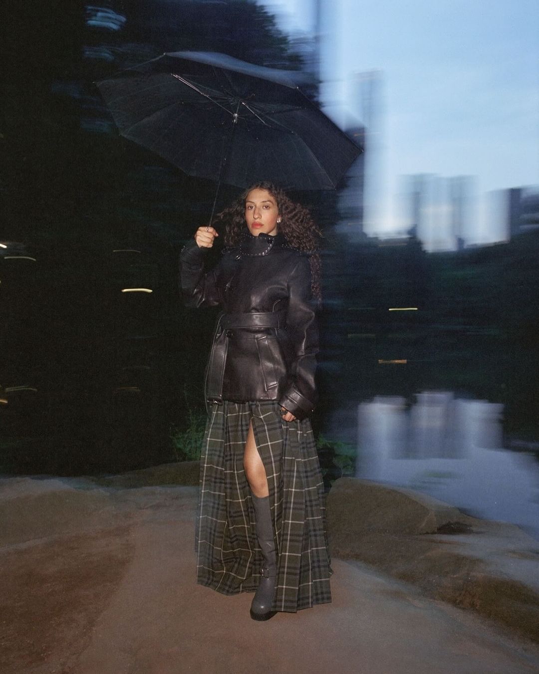 Woman stands in park at night holding umbrella. She wears a black leather jacket, plaid skirt, gray boots. Long exposure blurs background.