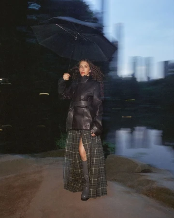 Woman stands in park at night holding umbrella. She wears a black leather jacket, plaid skirt, gray boots. Long exposure blurs background.