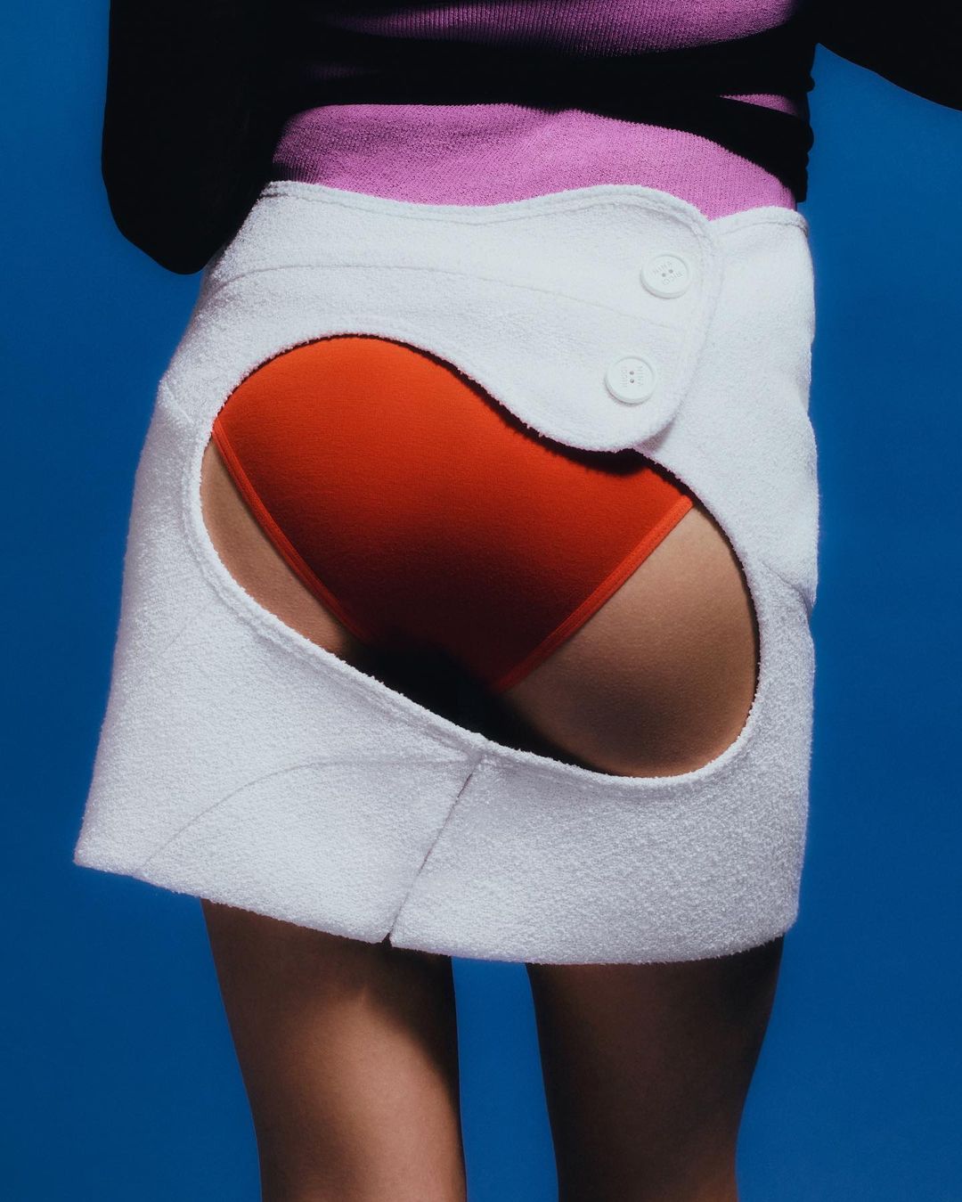 Model wearing a white cut-out skirt over red briefs and a pink top against a solid blue background by Leeor Wild.