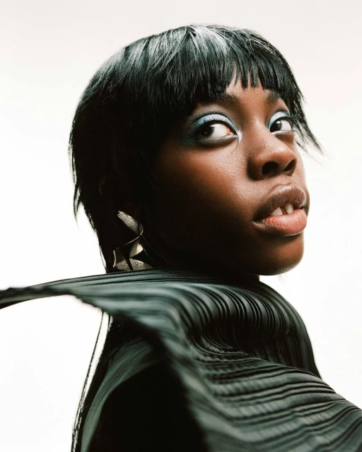 Portrait of an African-American woman with short black hair and blue eyeshadow, posing in front of a white background with jewelry.