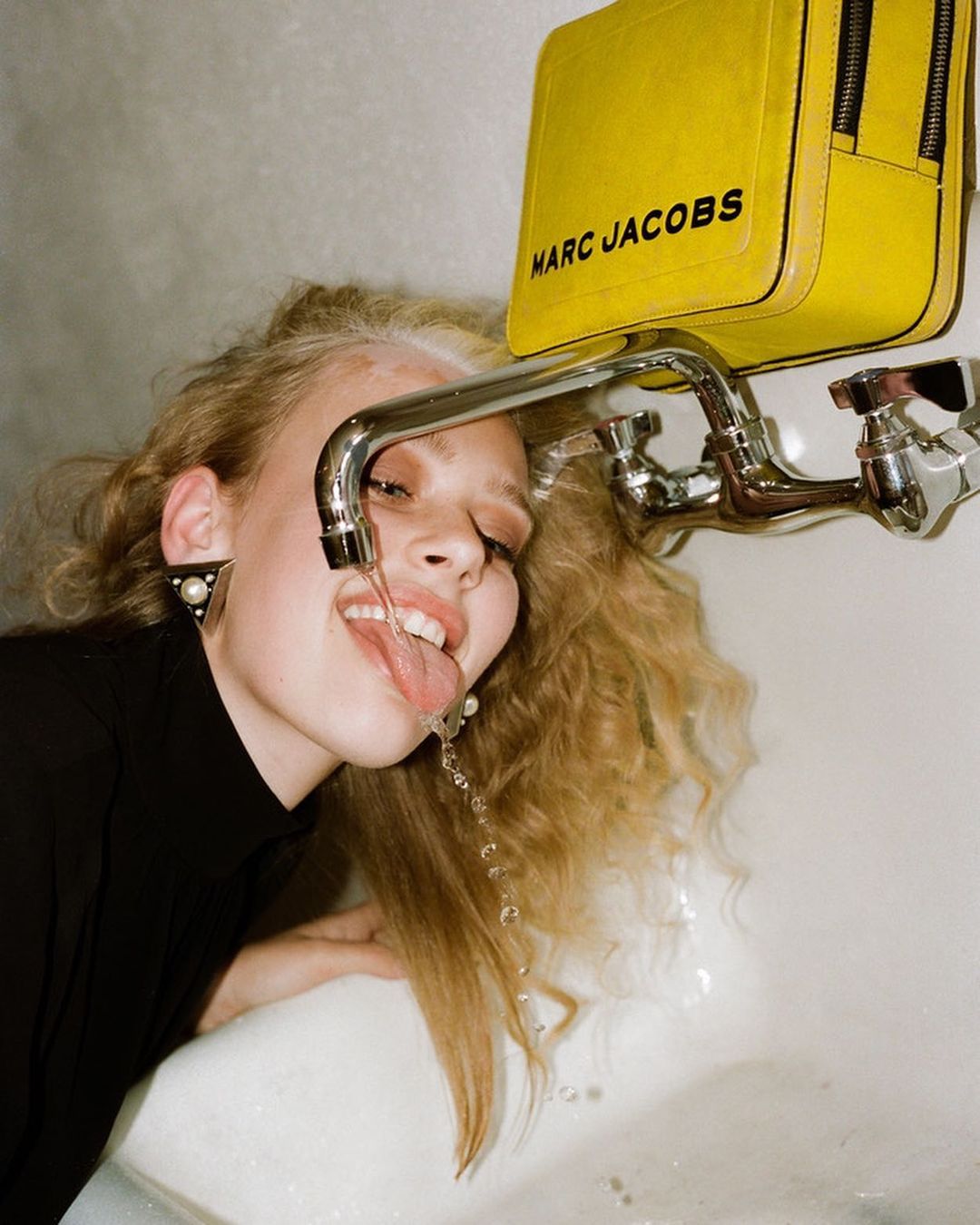 Young woman with blonde hair drinking water from a tap. She wears a black top, pearl earrings, and a yellow Marc Jacobs bag hangs above.