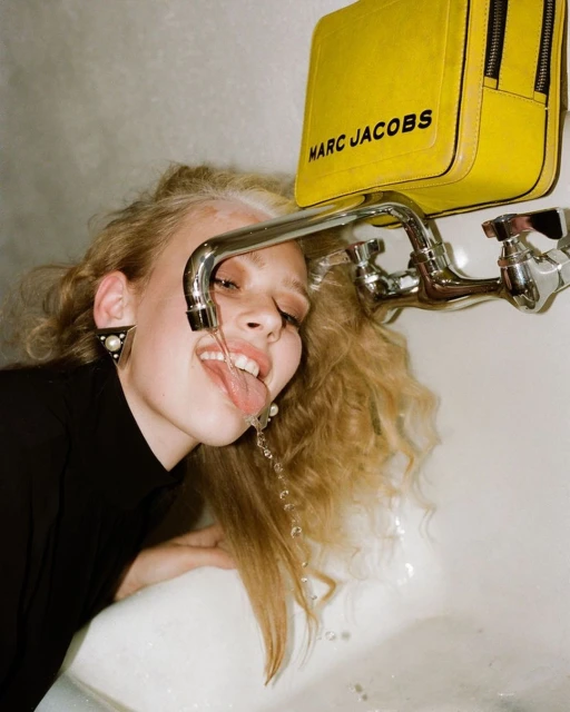 Young woman with blonde hair drinking water from a tap. She wears a black top, pearl earrings, and a yellow Marc Jacobs bag hangs above.