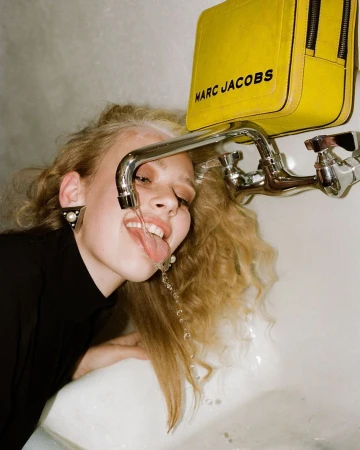 Young woman with blonde hair drinking water from a tap. She wears a black top, pearl earrings, and a yellow Marc Jacobs bag hangs above.
