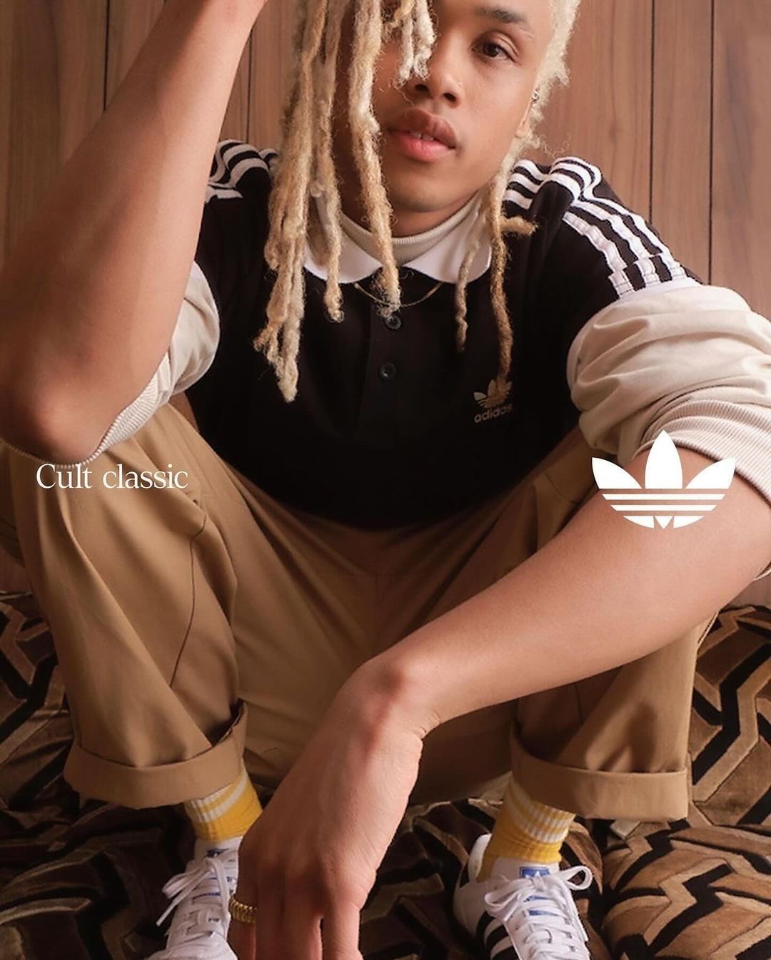 Young adult poses indoors wearing Adidas branded shirt, trousers, socks, and sneakers, with a patterned backdrop in a studio shot.