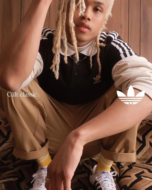 Young adult poses indoors wearing Adidas branded shirt, trousers, socks, and sneakers, with a patterned backdrop in a studio shot.