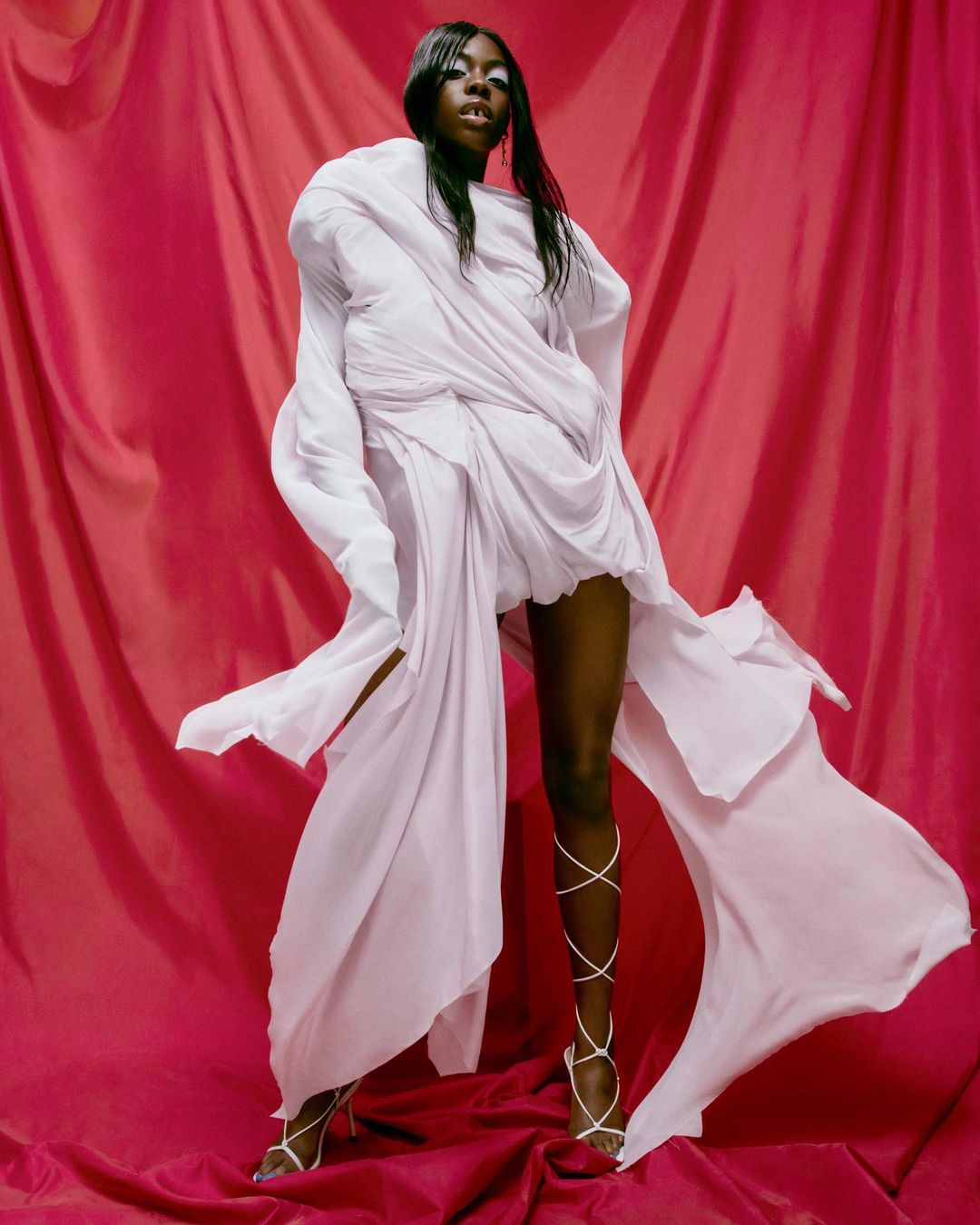 A Black woman poses in a light pink draped dress and gladiator heels against a matching pink fabric backdrop in a fashion-style shot.