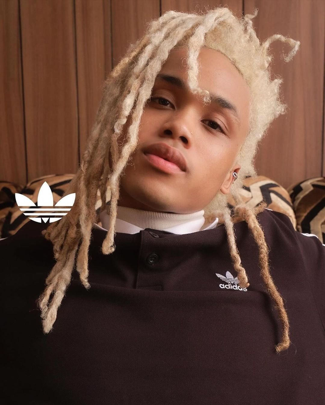 Portrait of a young adult male with blond dreadlocks, wearing a black and white Adidas shirt, posing indoors in front of a wooden wall.