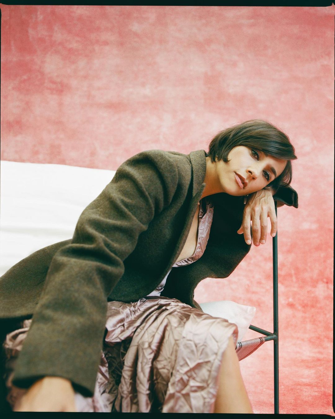 Adult woman in dark coat and light pink dress poses in front of light pink background in medium shot, resting hand on metal frame.