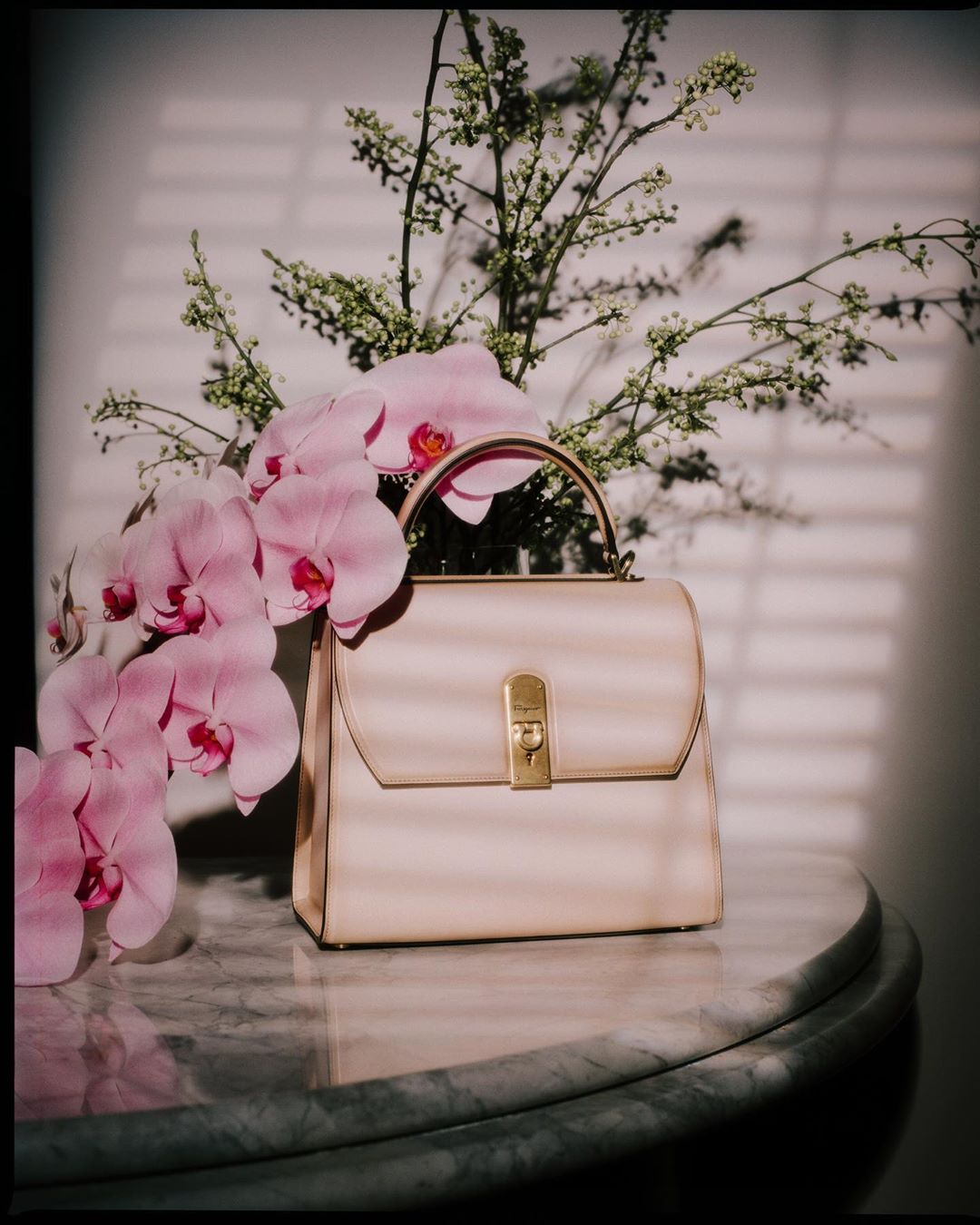 Elegant cream-colored purse with gold clasp, styled with pink orchids and greenery on a marble table, lit by window blinds.